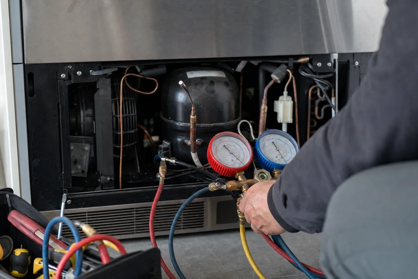 Manifold gauge set held at the compressor of a built-in refrigerator with the lower grille removed, open tool bag in front