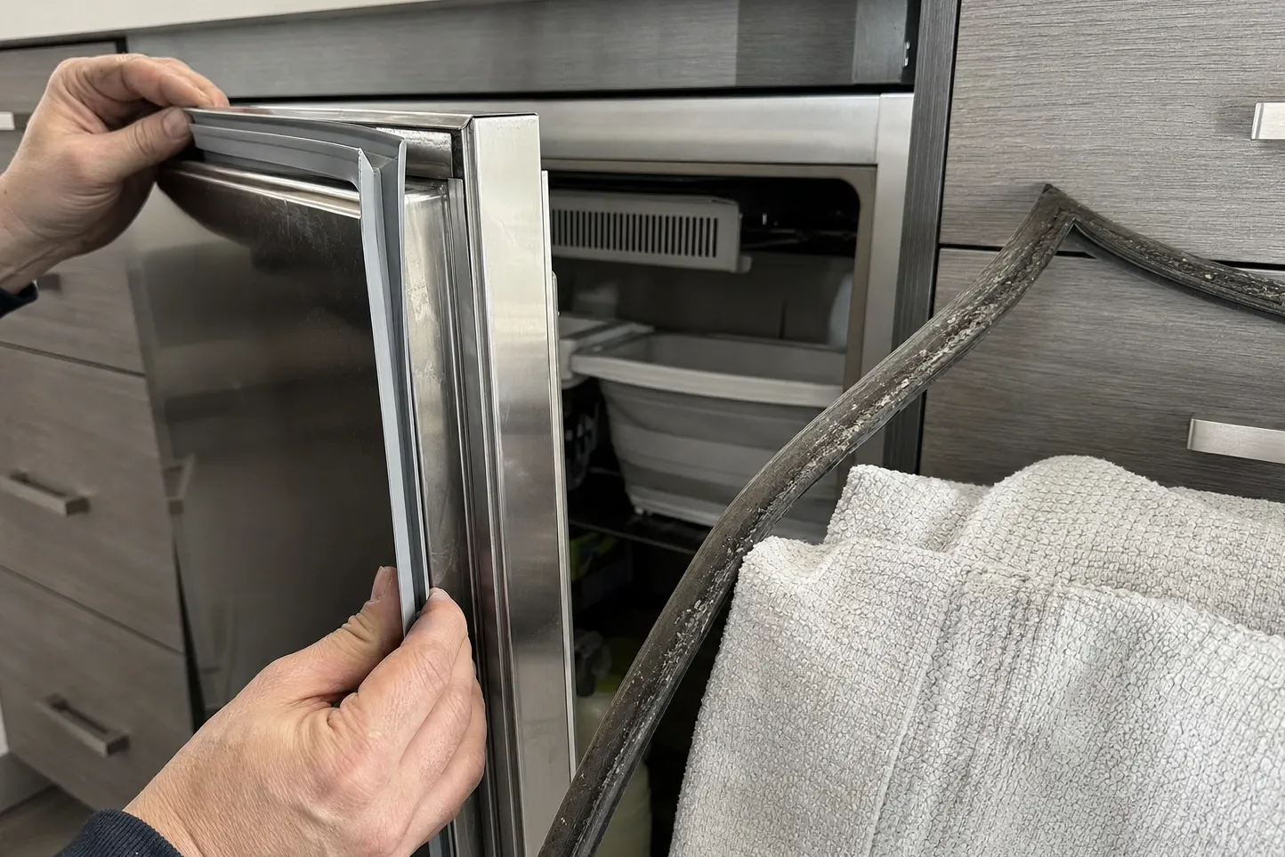 Hands holding a new grey door gasket against a stainless undercounter door, the blackened old gasket lying on a towel beside it